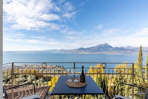 Une terrasse avec une table, deux verres à vin et une bouteille, donnant sur un lac et des montagnes.