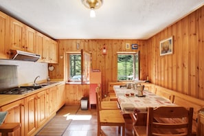 A cabin interior with wooden walls, a kitchen with a stove, a dining table with chairs, and a window with a view of trees.