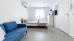 Una habitación compacta con cama, un sofá azul, una mesita con un vaso y un televisor en la pared.