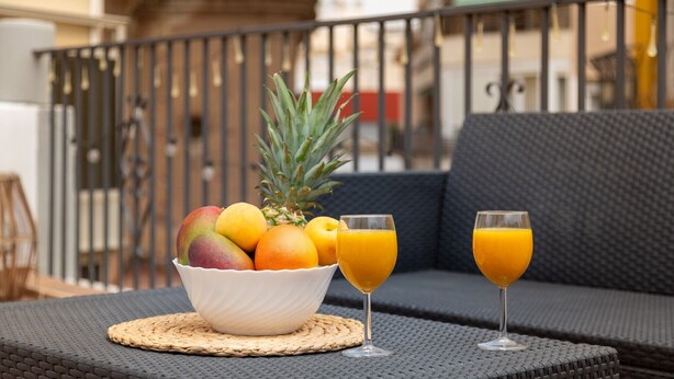 Un balcón con una mesa de mimbre, un cuenco de fruta y dos vasos de zumo de naranja.
