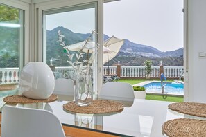 Un coin repas avec une table en verre, des chaises blanches, un vase transparent contenant des fleurs blanches, et une vue sur une piscine et des montagnes.