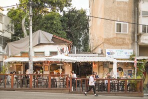 An outdoor cafe with tables and chairs, a covered seating area, and a building with a sign in the background. - Chic Red Studio near Talpiot Market (Haifa)