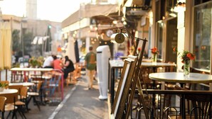 Um café ao ar livre com mesas e cadeiras, uma rua ao fundo e um edifício com um relógio.