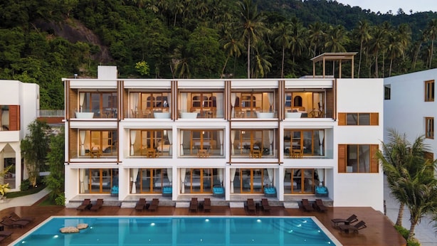 Un hotel moderno de varios pisos con una gran piscina, balcones con mobiliario y abundante vegetación al fondo.