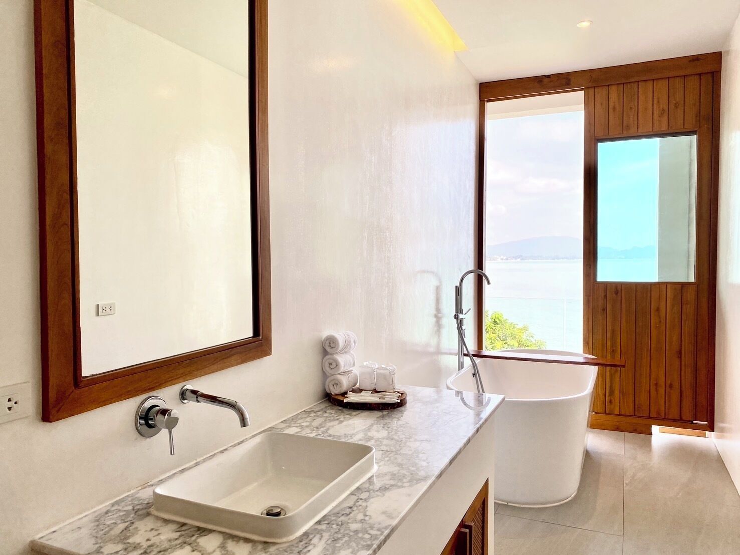 Ein modernes Badezimmer mit einem großen Spiegel, einem rechteckigen Waschbecken, einer freistehenden Badewanne und Blick auf ein Gewässer durch ein Fenster.