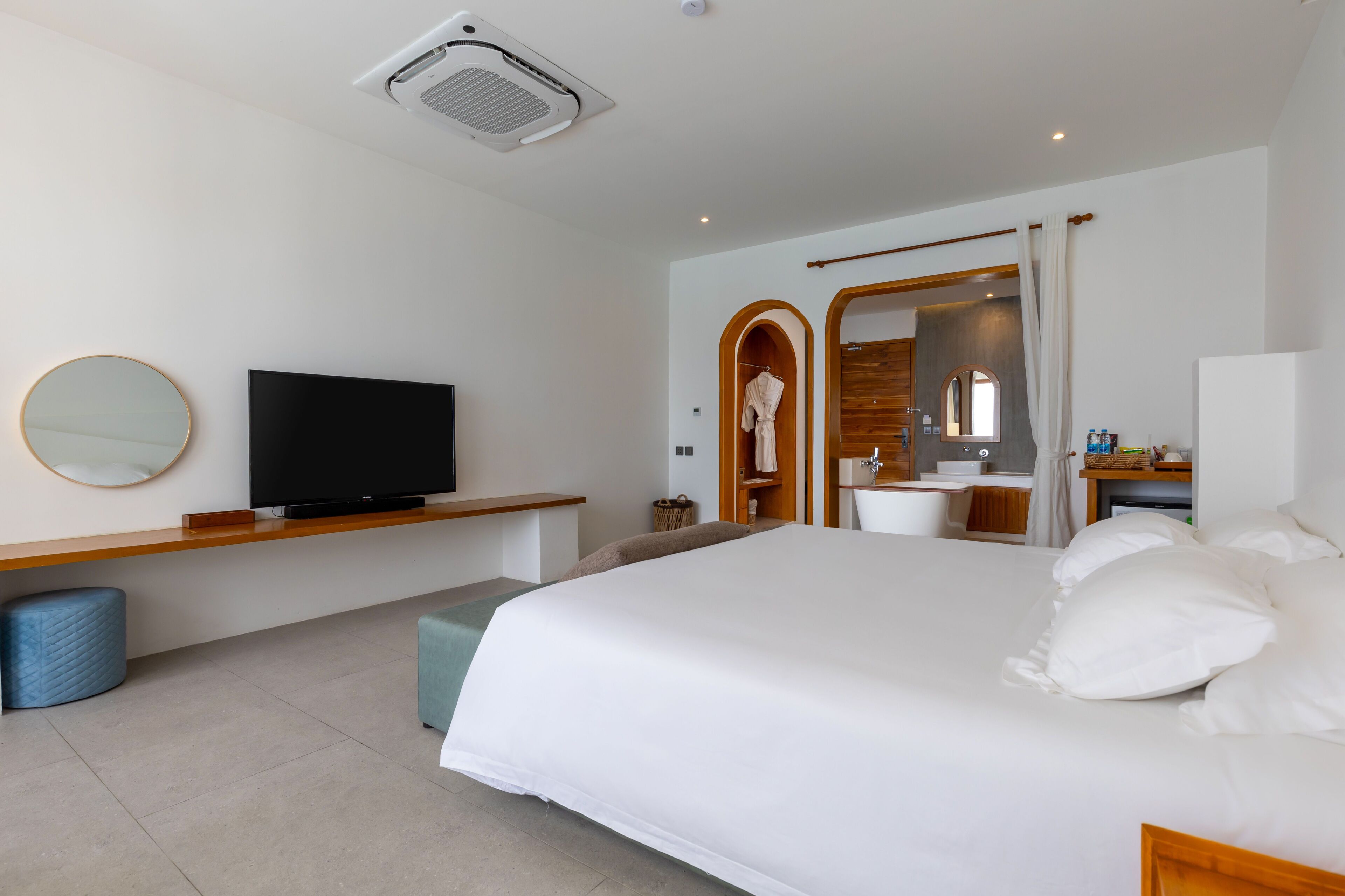 A modern hotel room with a large bed, a flat-screen TV, a wooden bedside table, a round mirror, and a bathroom with a wooden archway.