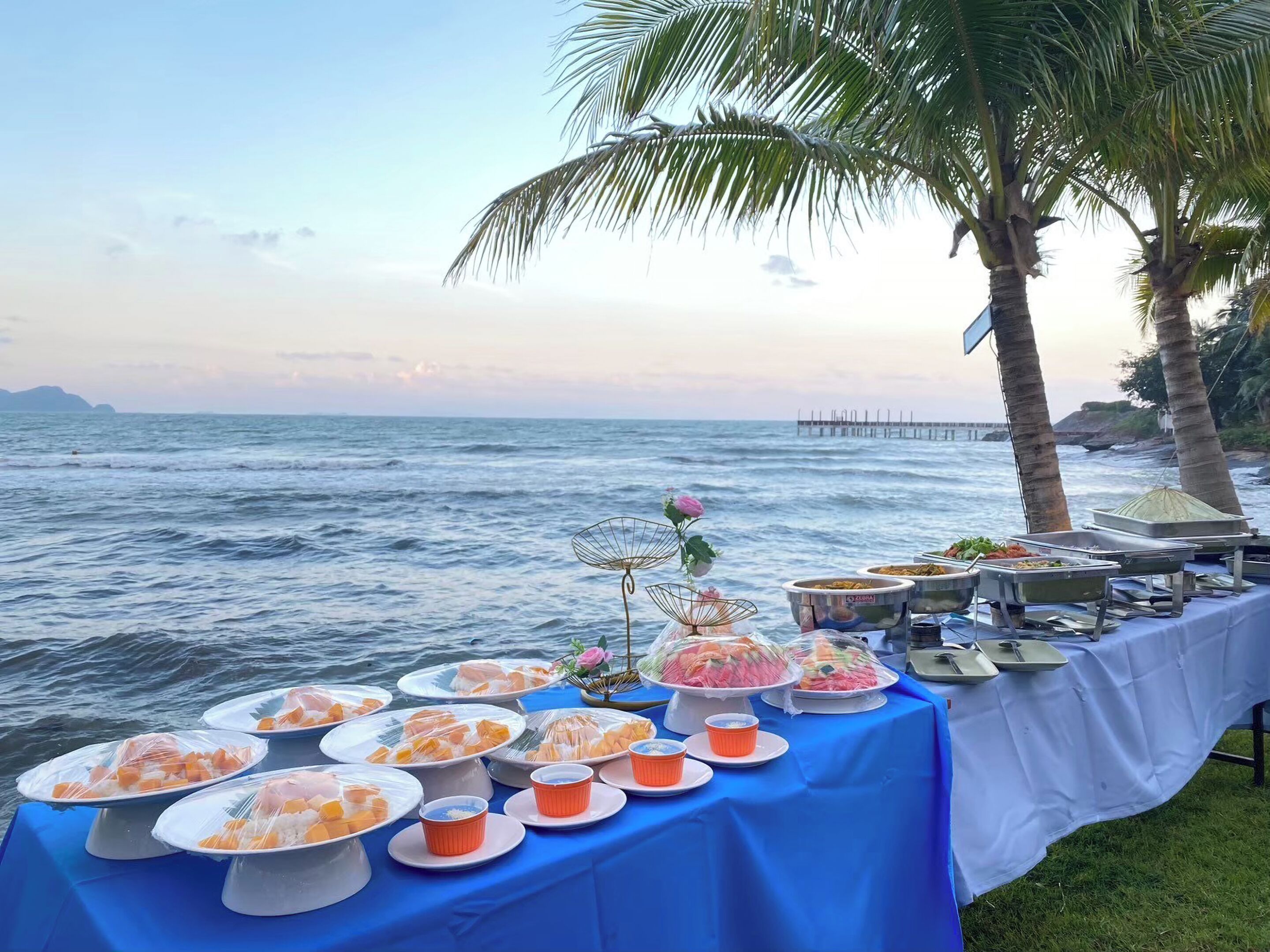 A buffet table with various dishes and a flower centerpiece, set against a coastal backdrop with palm trees and a pier.