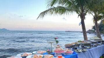 A buffet table with various dishes and a flower centerpiece, set against a coastal backdrop with palm trees and a pier.