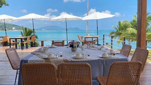 Comedor con vistas al océano, con sillas de mimbre, una mesa preparada para cuatro personas y sombrillas blancas.