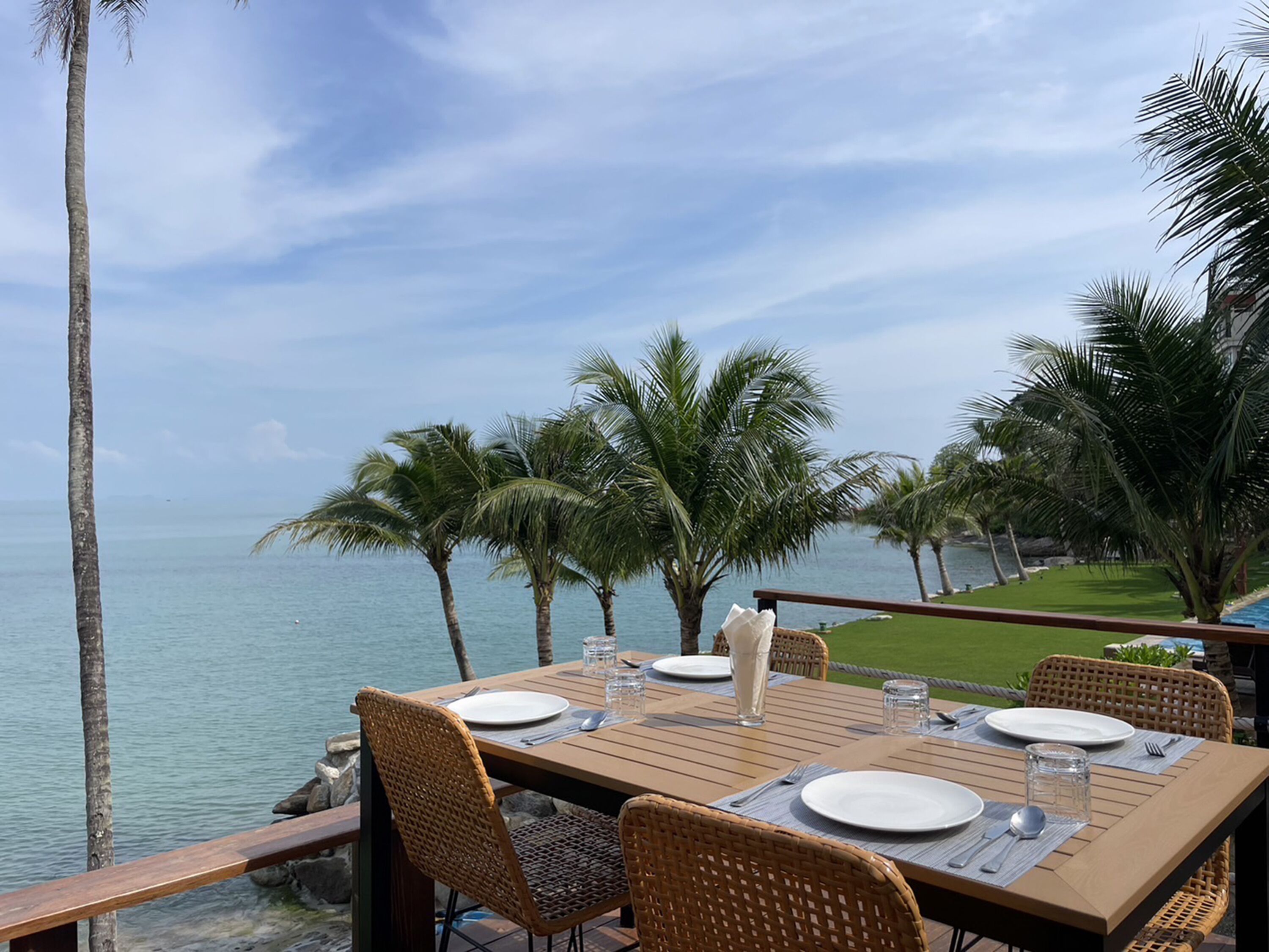 A dining table set with plates, cutlery, and glasses, overlooking a body of water with palm trees in the background.