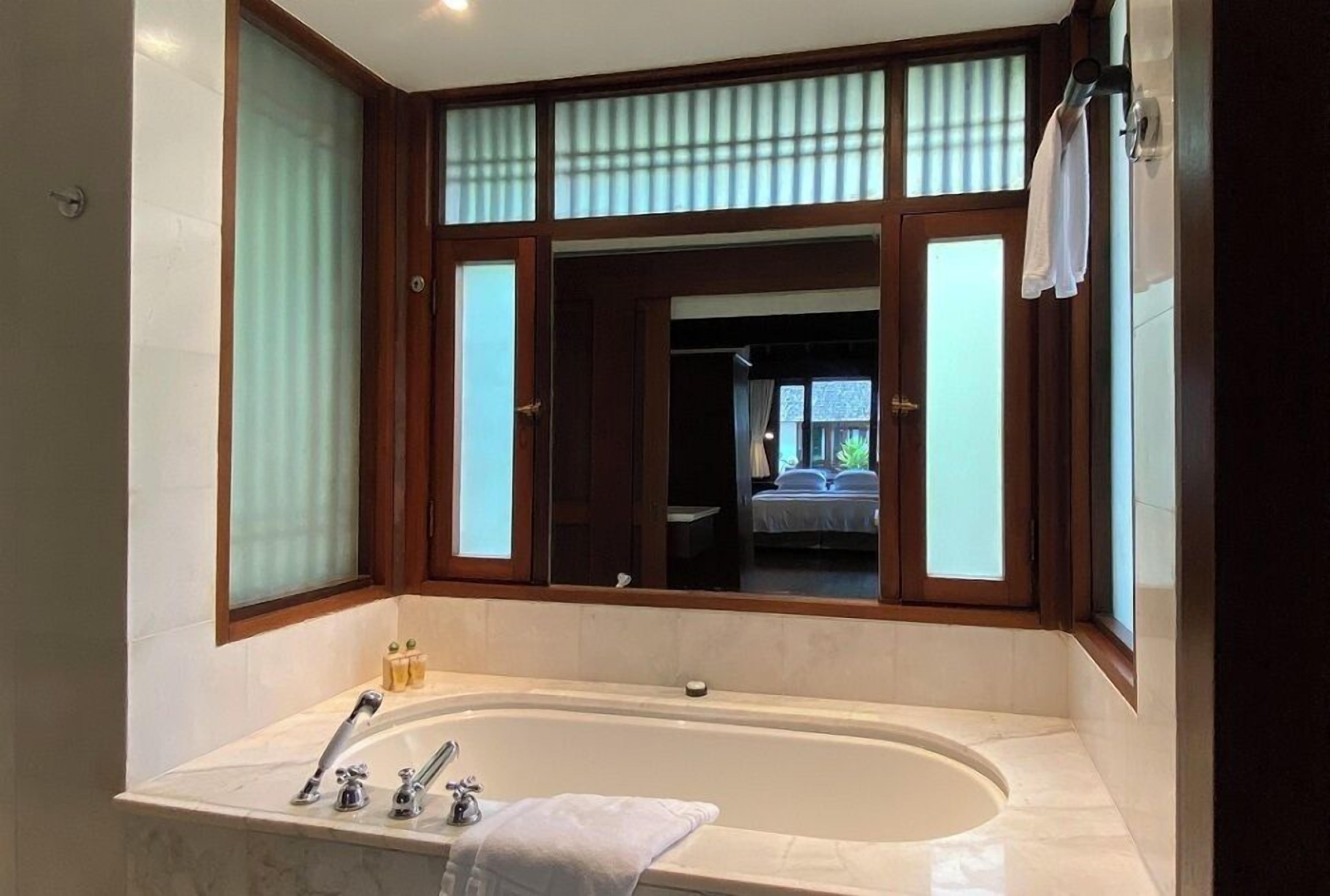 A bathroom with a large bathtub, a window with wooden frames, and a towel hanging on the wall.