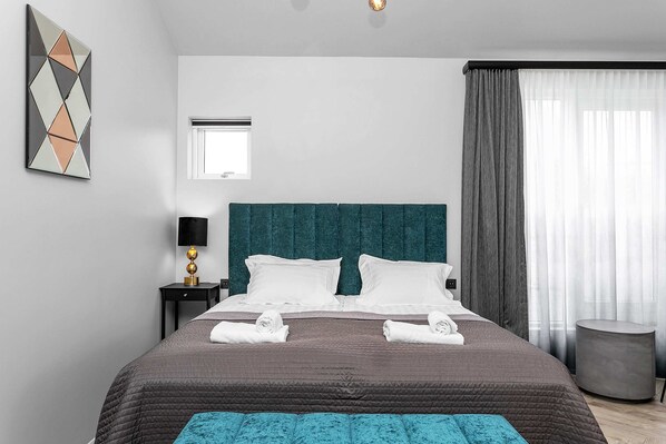A modern hotel room with a large bed, a headboard, a bedside table with a lamp, a round side table, and a window with curtains. - Backyard Stylish Studio for 2 - Excellent Location (Reykjavík)