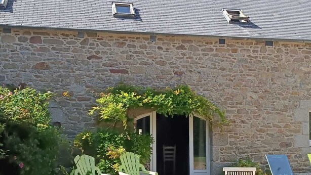A stone building with a tiled roof, a small garden with purple flowers, and outdoor seating. - Maison Dans Propriété Arborée, à 800m de la Plage (Plounéour-Brignogan-plages)