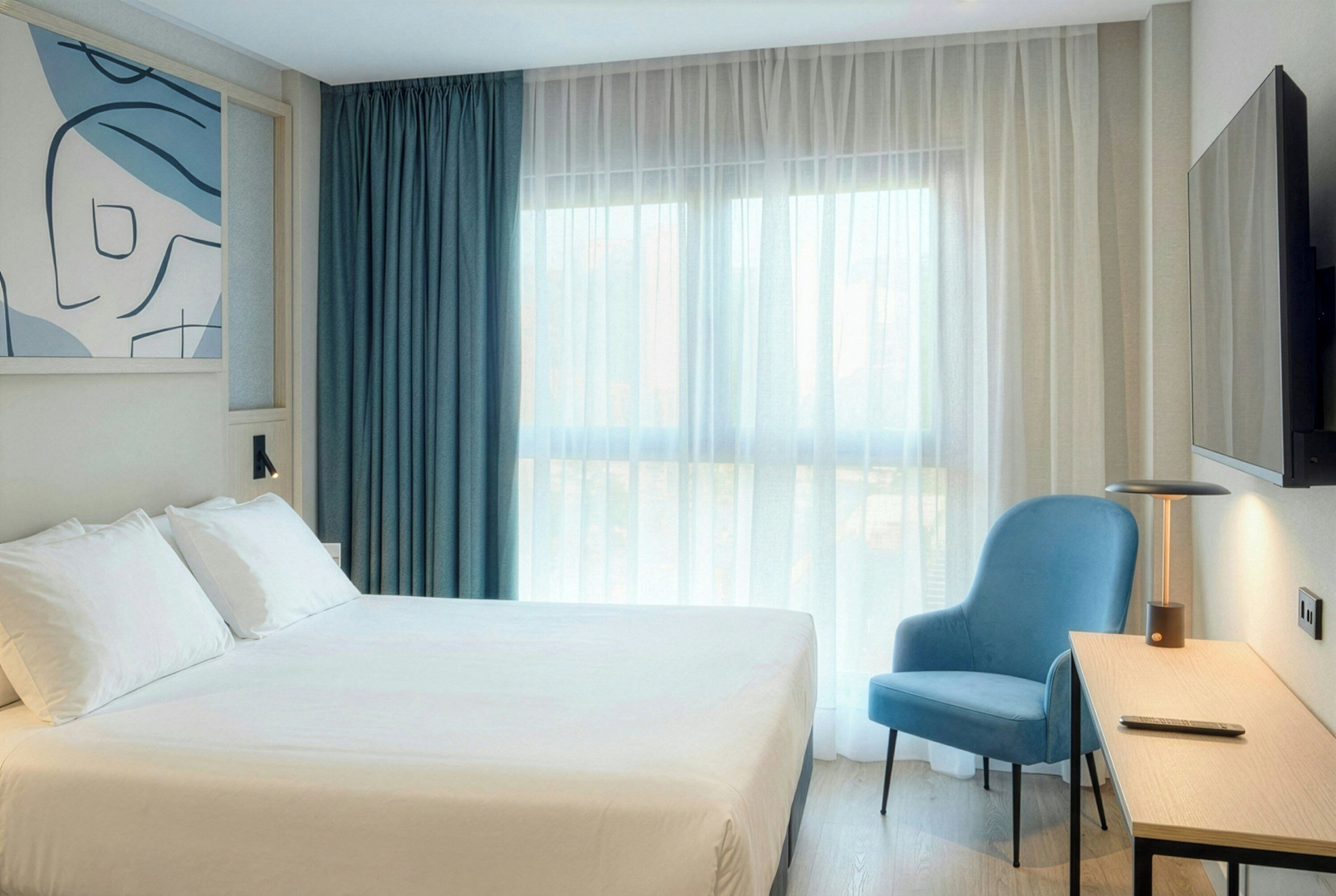 Una camera d'albergo con un letto, una sedia, una scrivania e un'ampia finestra con tende.