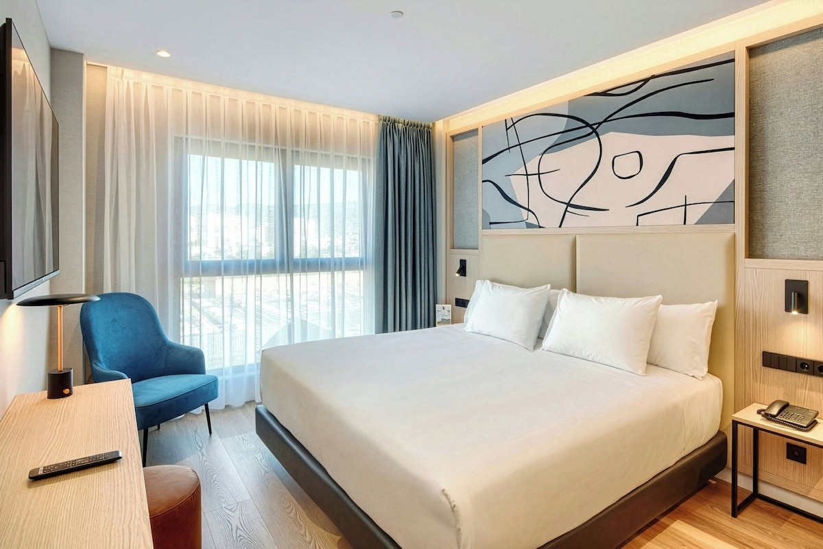 Una habitación de hotel con una cama grande, un escritorio con un control remoto, una silla, una mesita de noche con un teléfono y una ventana con cortinas.
