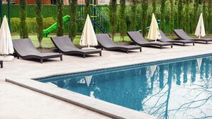 A poolside area with lounge chairs and umbrellas, a clear pool, and a green slide in the background. - Next Green Aparthotel Official (Batumi)