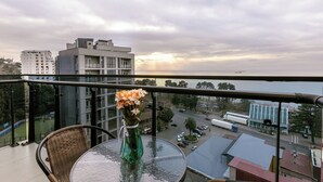 A balcony with a wicker chair, a glass table, and a vase of flowers. A cityscape with buildings and a body of water is visible in the background. - Next Green Aparthotel Official (Batumi)