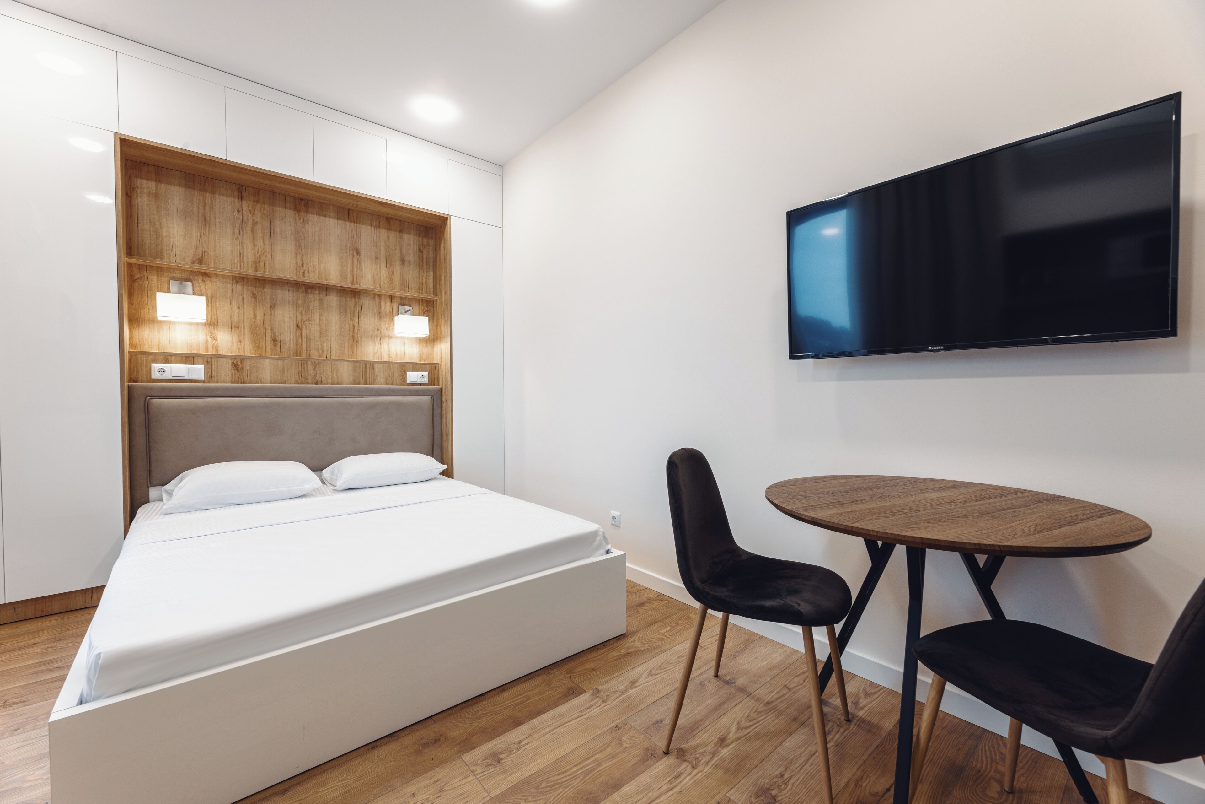 Moderni hotellihuone, jossa on sänky, seinälle asennettu televisio ja pieni pöytä tuoleineen.