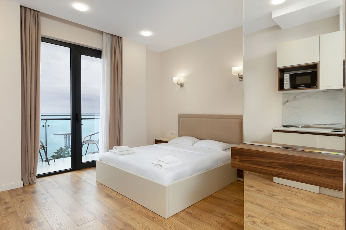 A modern hotel room with a large bed, a wooden desk, a built-in microwave, and a balcony with a view.