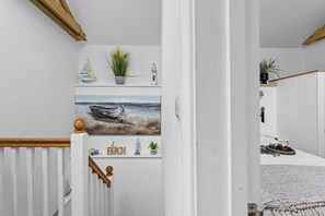 A hallway with a staircase, a painting of a boat on water, and a shelf with decorative items. - The Boathouse (Torquay)