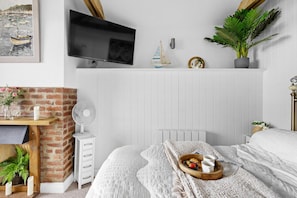 Ein Schlafzimmer mit Kamin aus Ziegelsteinen, einem Bett mit weißer, strukturierter Decke, einem kleinen Ventilator und einer Holztablett mit Tassen und einer Schüssel.