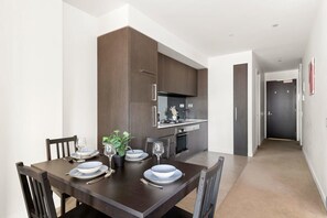 A modern dining area with a dark wooden table set for four, a kitchen with dark cabinetry, and a hallway leading to a door. - Stylish & Modern 2BR Apt Near Victoria Market (Melbourne)