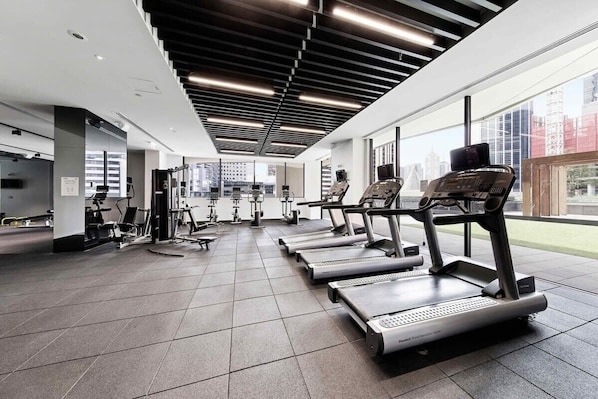 Une salle de sport moderne équipée de tapis de course, d’appareils de musculation et dotée d’une grande fenêtre offrant une vue sur la ville.