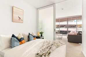 A modern bedroom with a large bed, a bedside table with a lamp, and a view of a balcony with a sofa. - Stylish & Modern 2BR Apt Near Victoria Market (Melbourne)