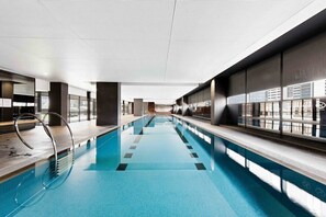 A modern indoor swimming pool with clear glass walls, a long poolside area, and a view of the cityscape. - Stylish & Modern 2BR Apt Near Victoria Market (Melbourne)