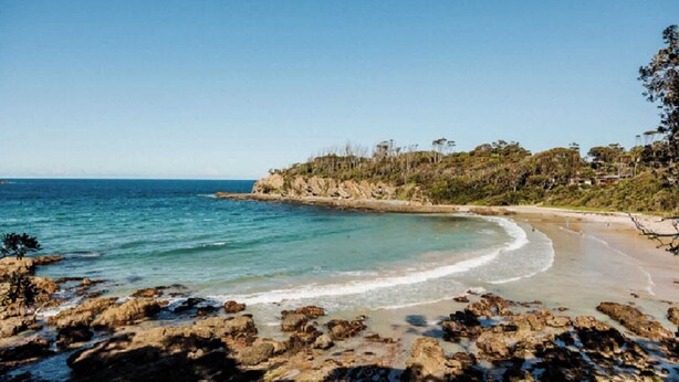 A coastal beach with rocky foreground, clear blue water, and a forested hillside in the background. - Pet-Friendly, WiFi & Pool Table | Two Salty Dogs (Lilli Pilli)