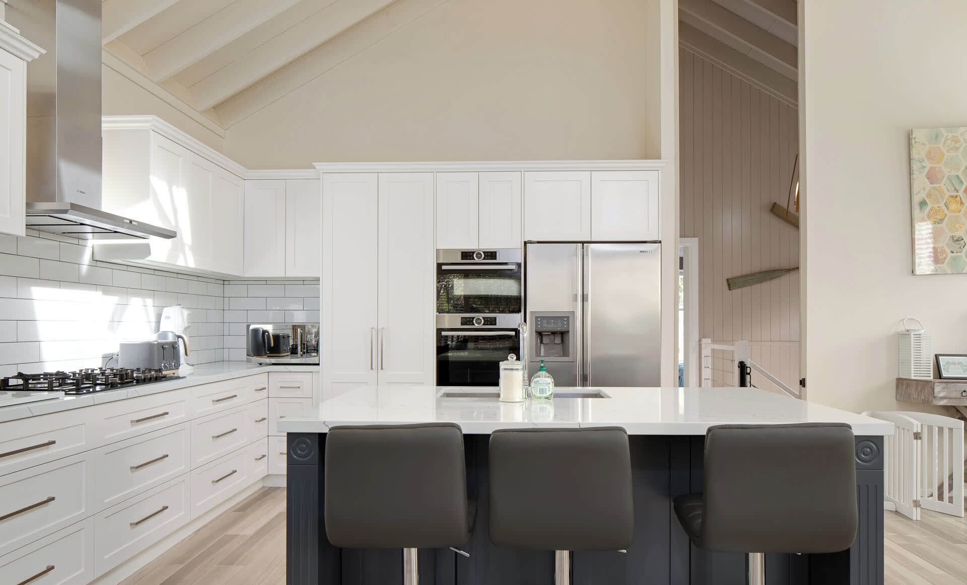 Una cocina moderna con muebles blancos, una isla central con taburetes altos, electrodomésticos de acero inoxidable y un techo alto con vigas a la vista.