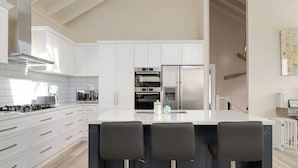 A modern kitchen with white cabinetry, a central island with bar stools, stainless steel appliances, and a high ceiling with exposed beams. - Pet-Friendly, WiFi & Pool Table | Two Salty Dogs (Lilli Pilli)