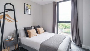 Une chambre à coucher comprenant un lit, une table de chevet, un porte-manteau mural, une fenêtre avec des rideaux et un tableau encadré accroché au mur.