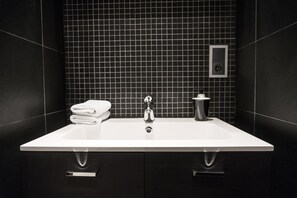 A modern bathroom with a large white sink, black tiled walls, and a small speaker mounted on the wall.