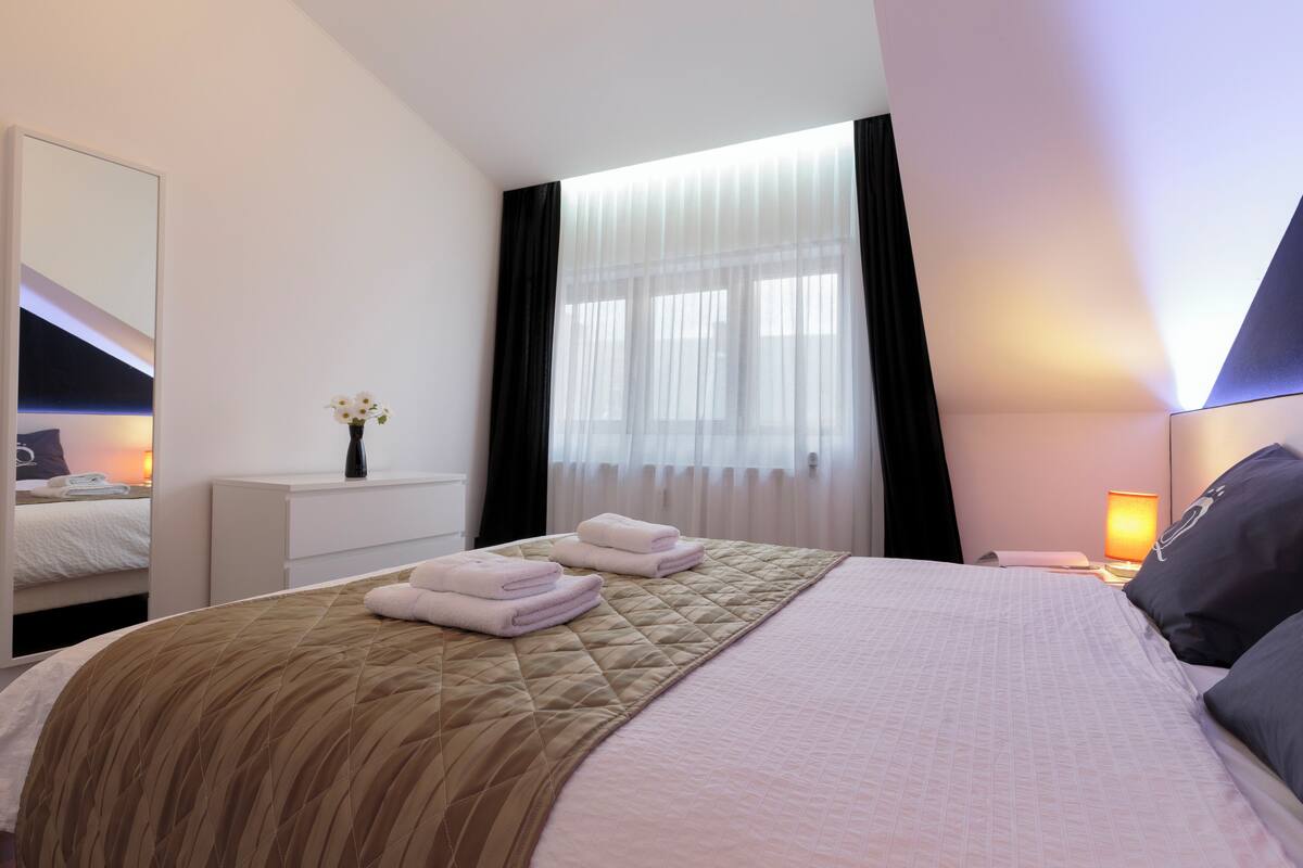 Ein Hotelzimmer mit einem großen Bett, einem Nachttisch mit Lampe, einem Spiegel und einem Fenster mit durchscheinenden Vorhängen.