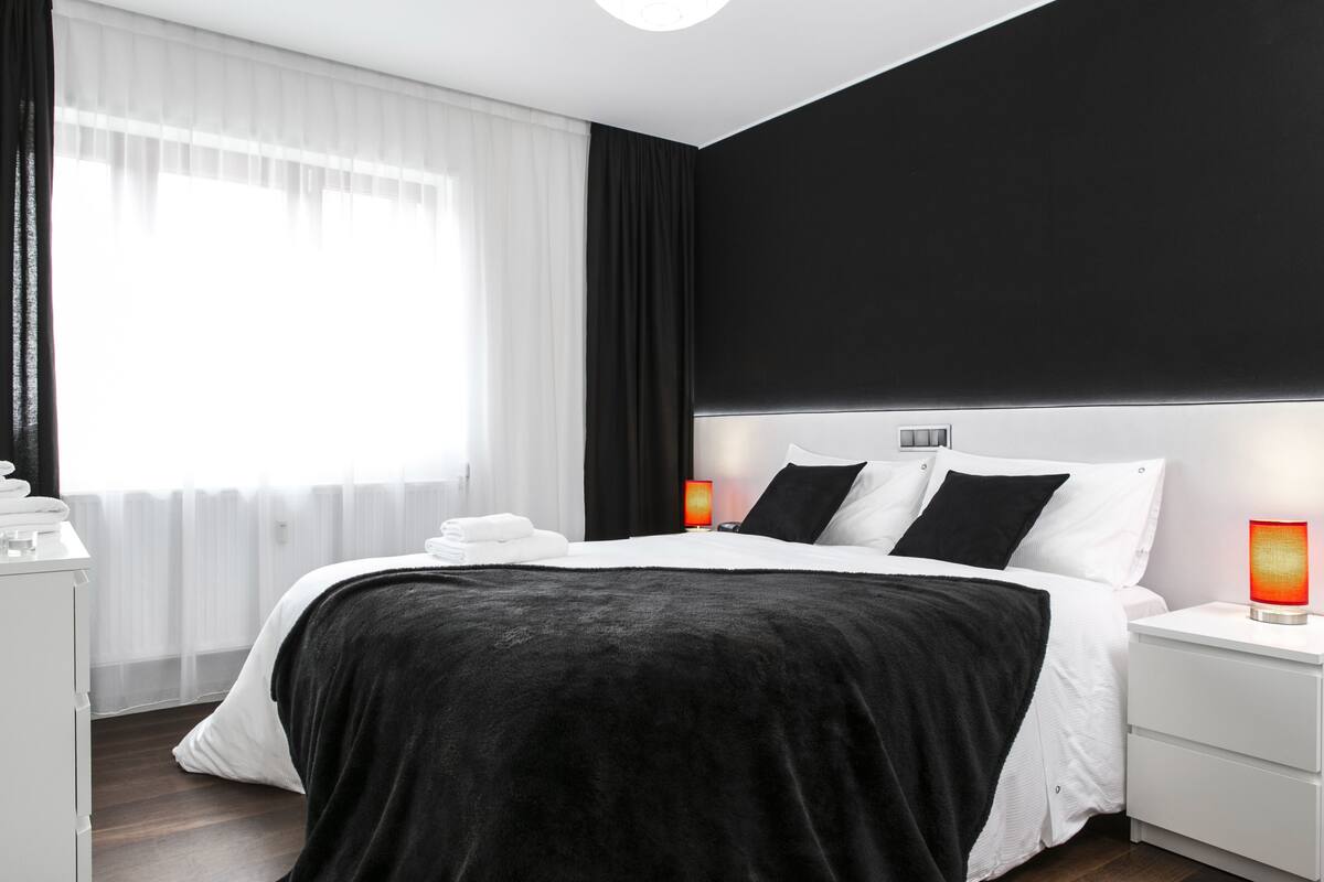Ein modernes Schlafzimmer mit einem großen Bett, weißen Nachttischen, einem schwarzen Kopfteil und einem Fenster mit durchsichtigen Vorhängen.