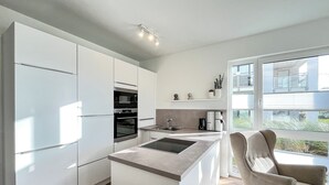 Una cocina moderna con armarios blancos, horno empotrado, microondas y cafetera. Hay un ventanal con vistas al exterior.