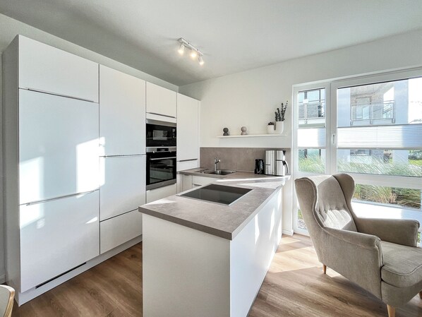 Una cocina moderna con armarios blancos, horno empotrado, microondas y cafetera. Hay un ventanal con vistas al exterior.