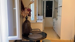 A compact living space with a dining area featuring a table and chairs, a small stool, and a vase with flowers. A kitchen with a refrigerator and stove is visible in the background. - THE CENTRIC Apt. by LOC HOSPITALITY (Corfu)