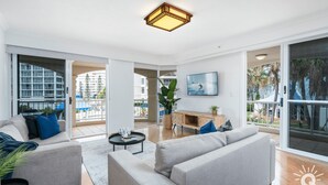 A modern living room with a grey sofa set, a coffee table, and a TV mounted on the wall. There is a balcony with a view of the city and palm trees. - The Moroccan - 3 Bedroom Beachside Unit - Q Stay (Surfers Paradise)