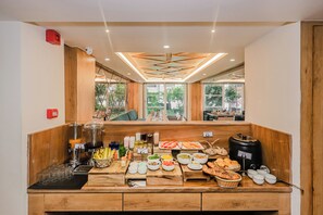 A breakfast buffet with various food items and beverages, wooden paneling, and large windows with a view of greenery.