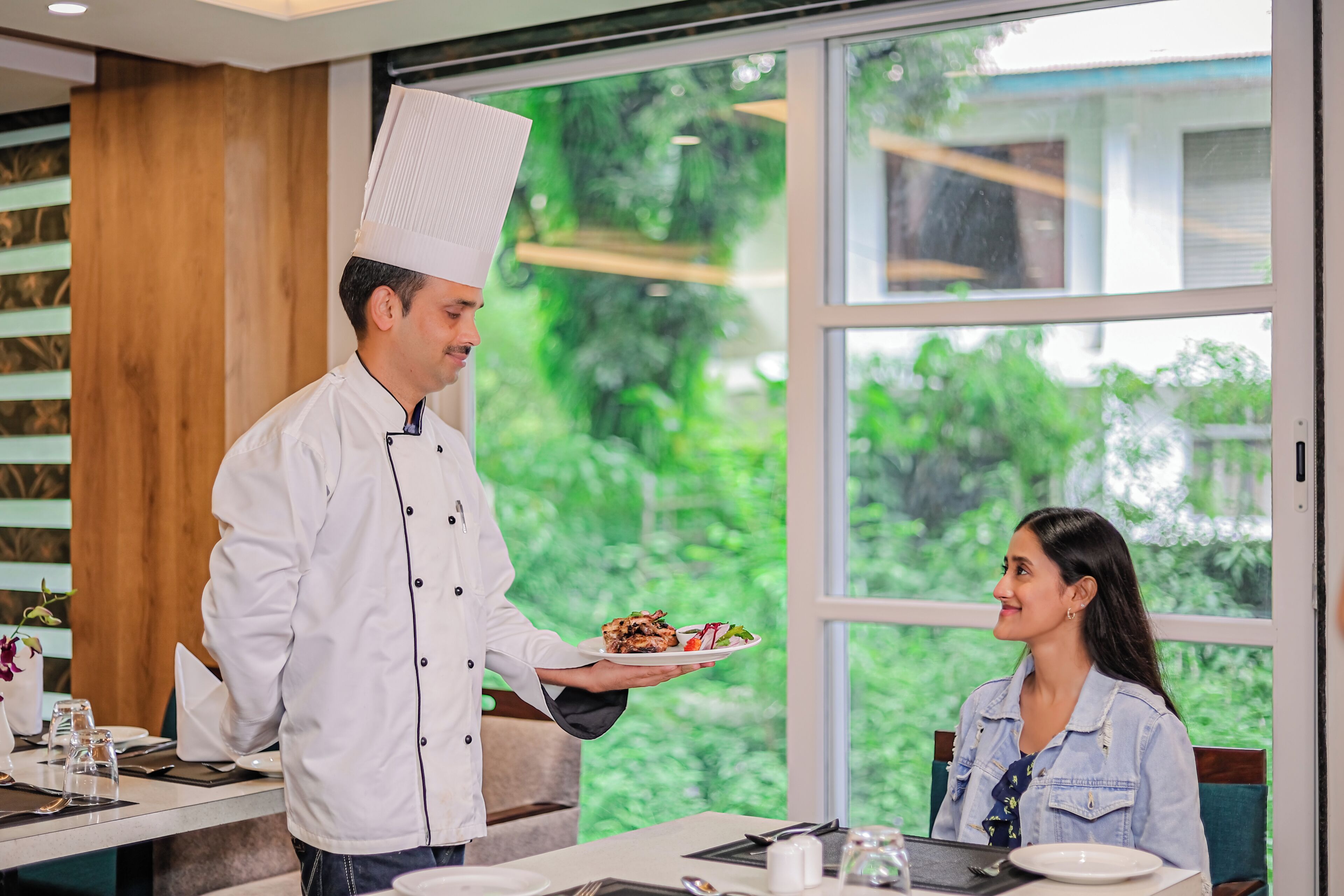 A chef serving food to a guest in a restaurant with large windows and a view of greenery.