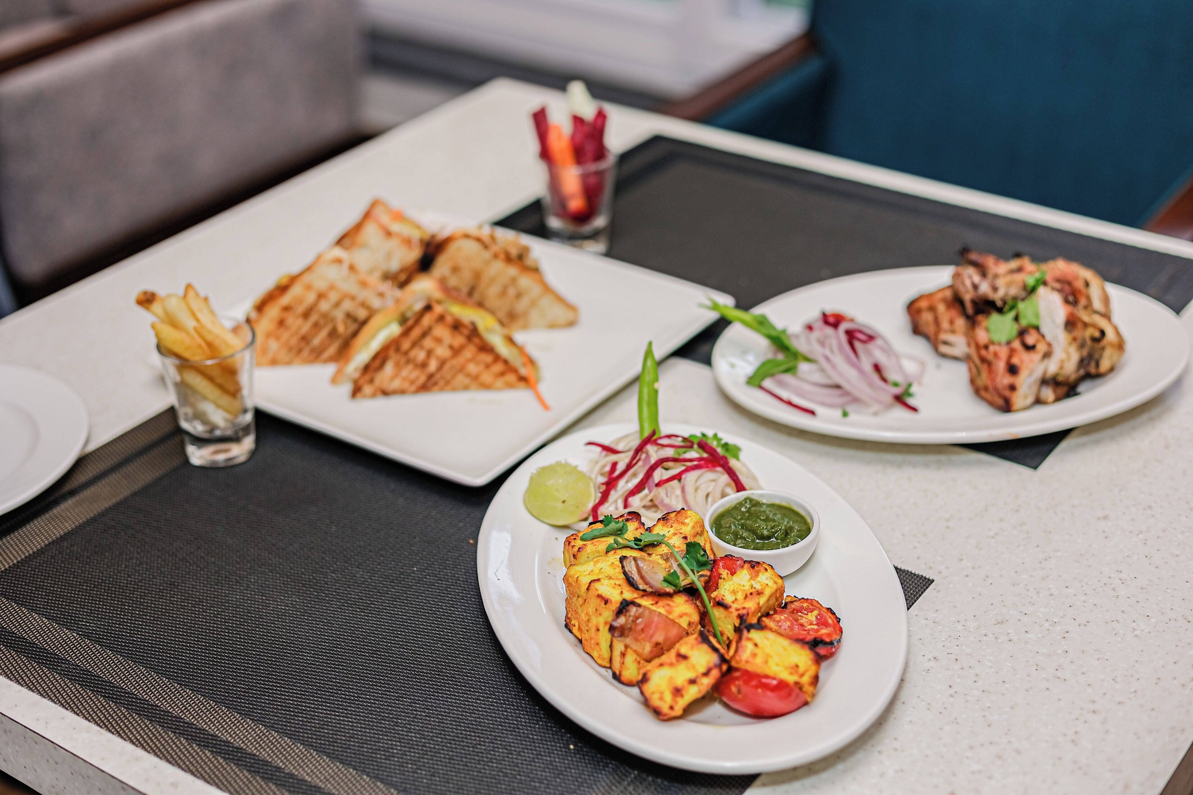 A table with a variety of dishes including grilled chicken, paneer, and sandwiches, accompanied by condiments and drinks.
