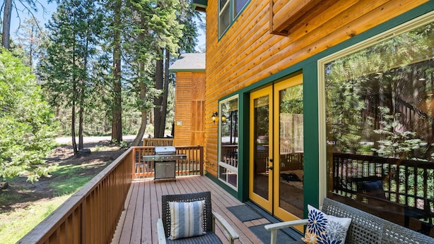 Une terrasse en bois avec des meubles d’extérieur, un barbecue et une grande fenêtre donnant sur une zone boisée.