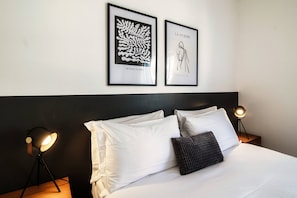 A neatly made bed with white linens and a dark pillow, flanked by two bedside lamps. Above the bed, two framed artworks are hung on the wall. - A Casa di Laura (Lugano)