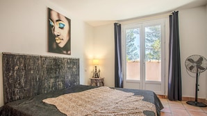 A bedroom with a large bed, a headboard, a nightstand with a lamp, a window with curtains, and a standing fan. - Beautiful home in Les Arcs (Les Arcs)