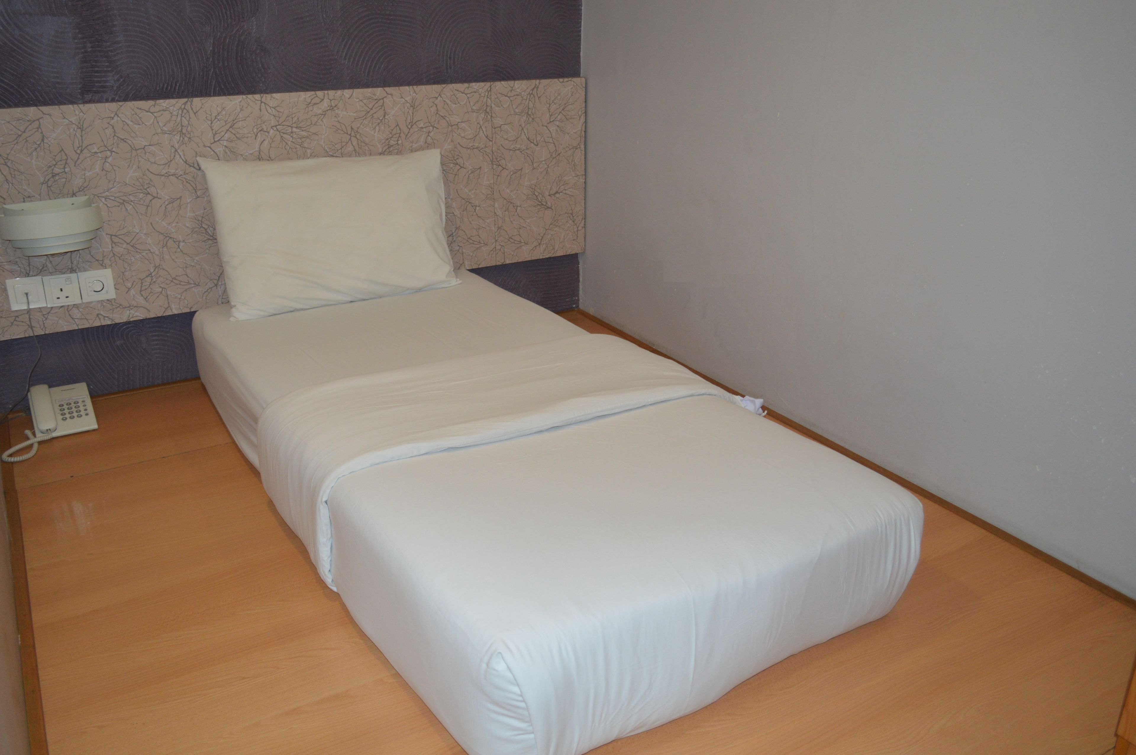 A single bed with a white mattress and pillow, a bedside table with a white phone, and a wall socket.