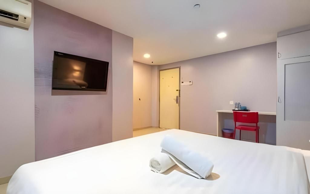 A hotel room with a bed, a TV mounted on the wall, a desk with a chair, and a door leading to another room.