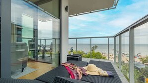 Un balcón con vista a la playa, una mesa con un sombrero y un libro, y un vidrio que da a un espacio interior.