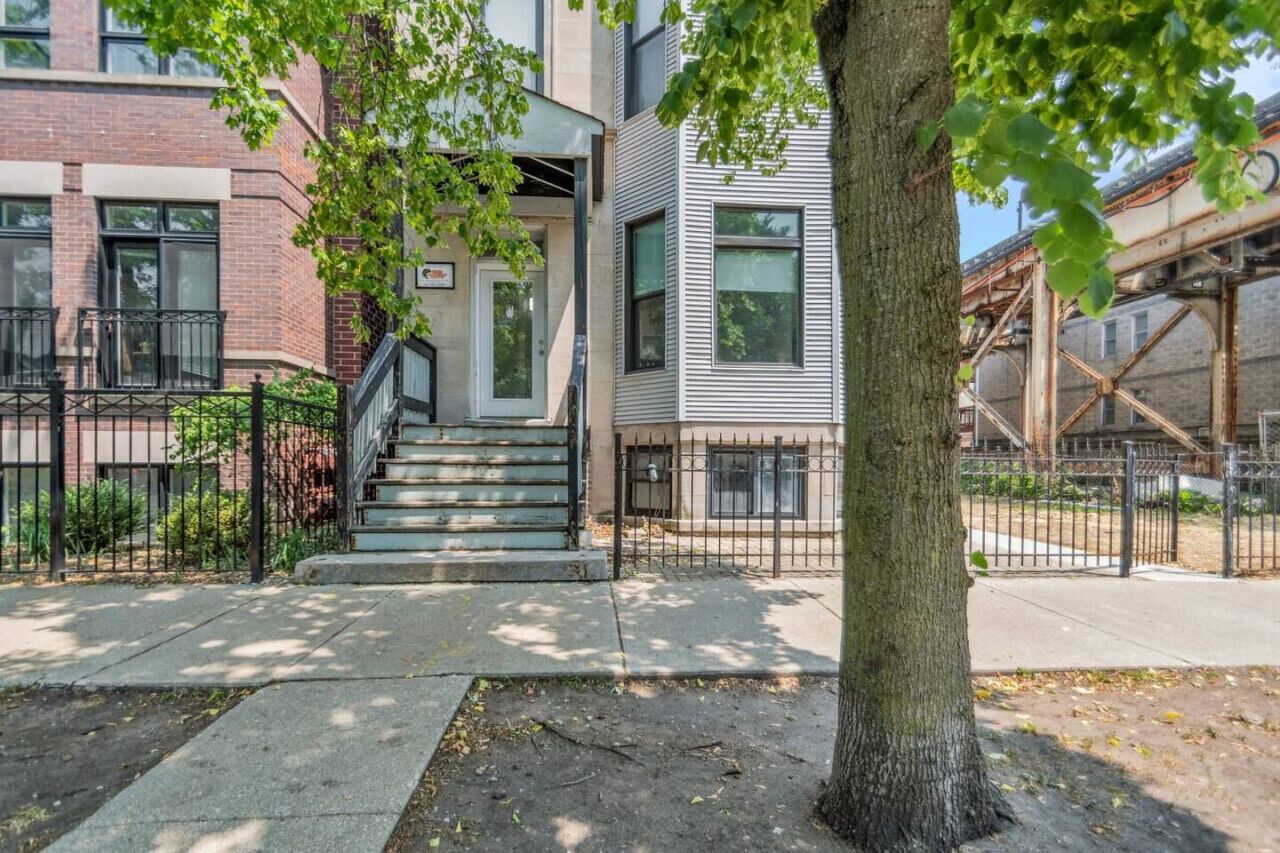 A residential building with a staircase leading to the entrance, a tree in the foreground, and a metal fence.